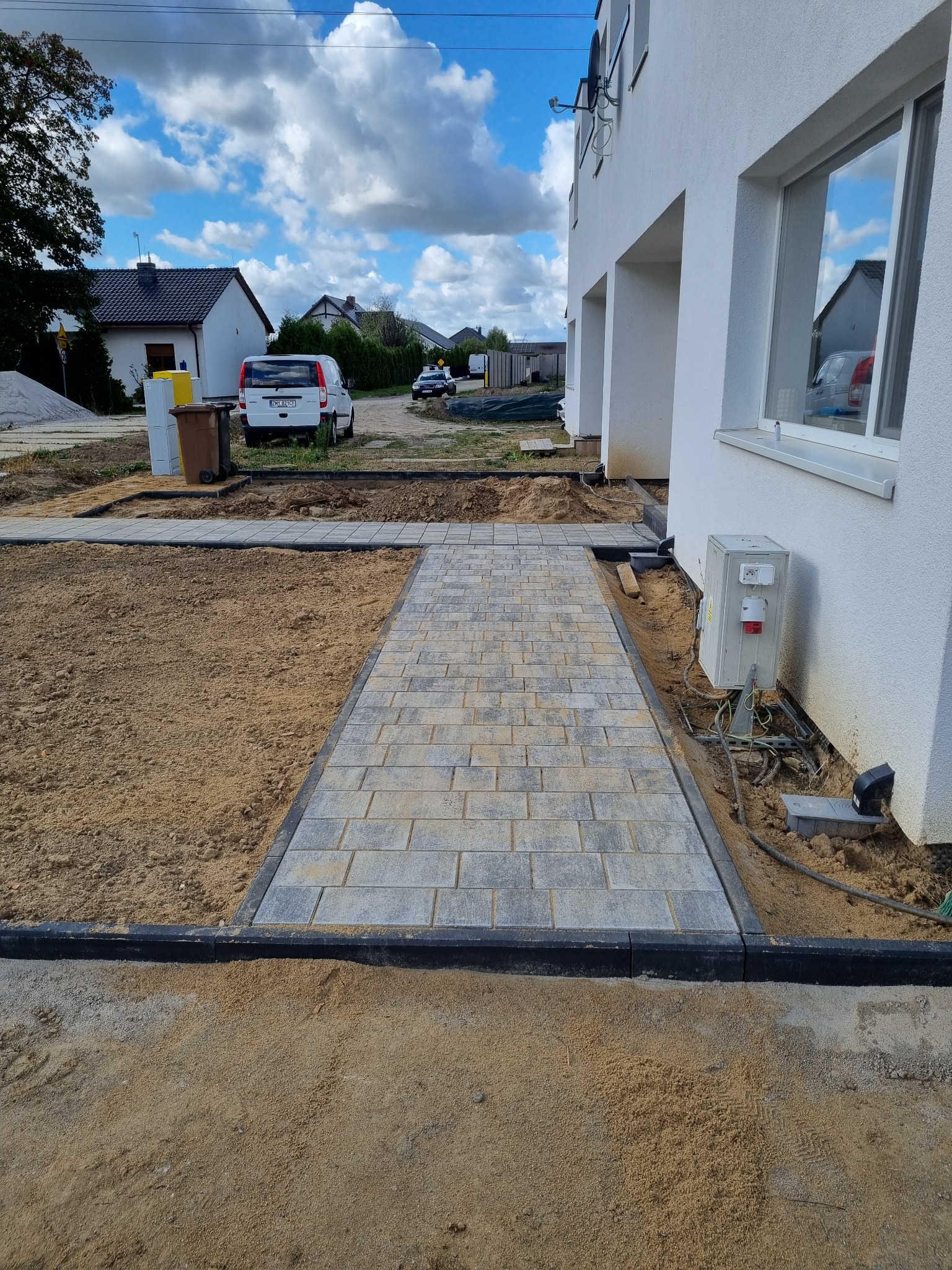 Ułożony chodnik z szarej kostki brukowej prowadzący do białego budynku, obrzeża wykończone czarnym krawężnikiem, widoczna ziemia przygotowana pod trawnik, na tle domy i błękitne niebo z chmurami.