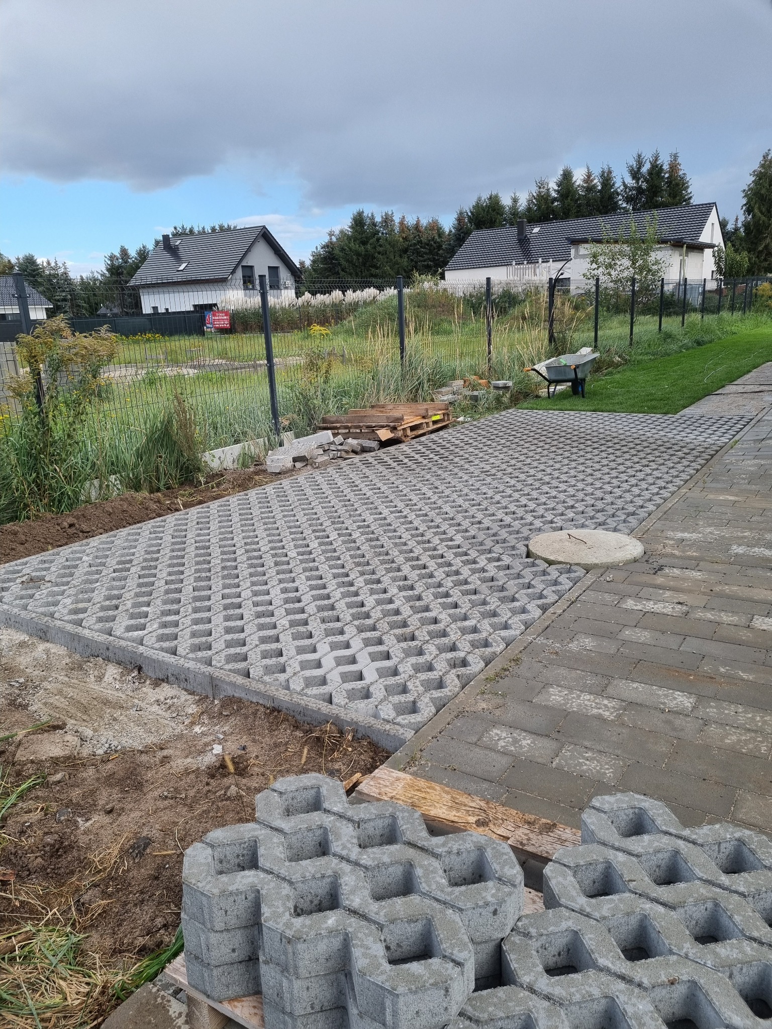 Ułożona nawierzchnia z ażurowych betonowych płyt na posesji, widoczne stosy kostki do dalszych prac, w tle dom jednorodzinny i ogrodzenie.