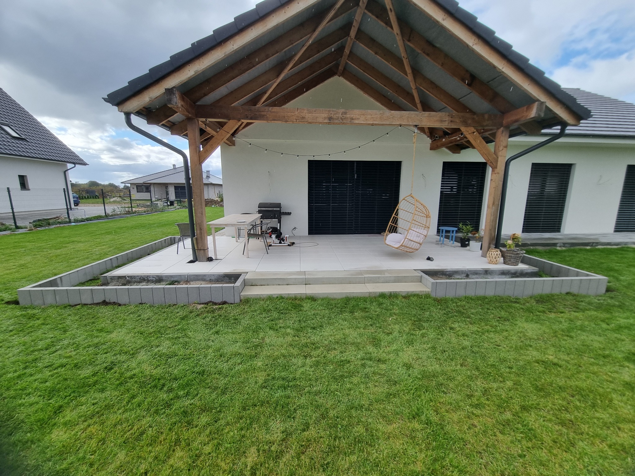 Nowoczesny taras z drewnianą konstrukcją zadaszenia, wykończony jasnymi płytami, otoczony obrzeżem z betonowych bloczków i trawnikiem. Widoczne elementy wyposażenia: wiszące krzesło, grill, stół...