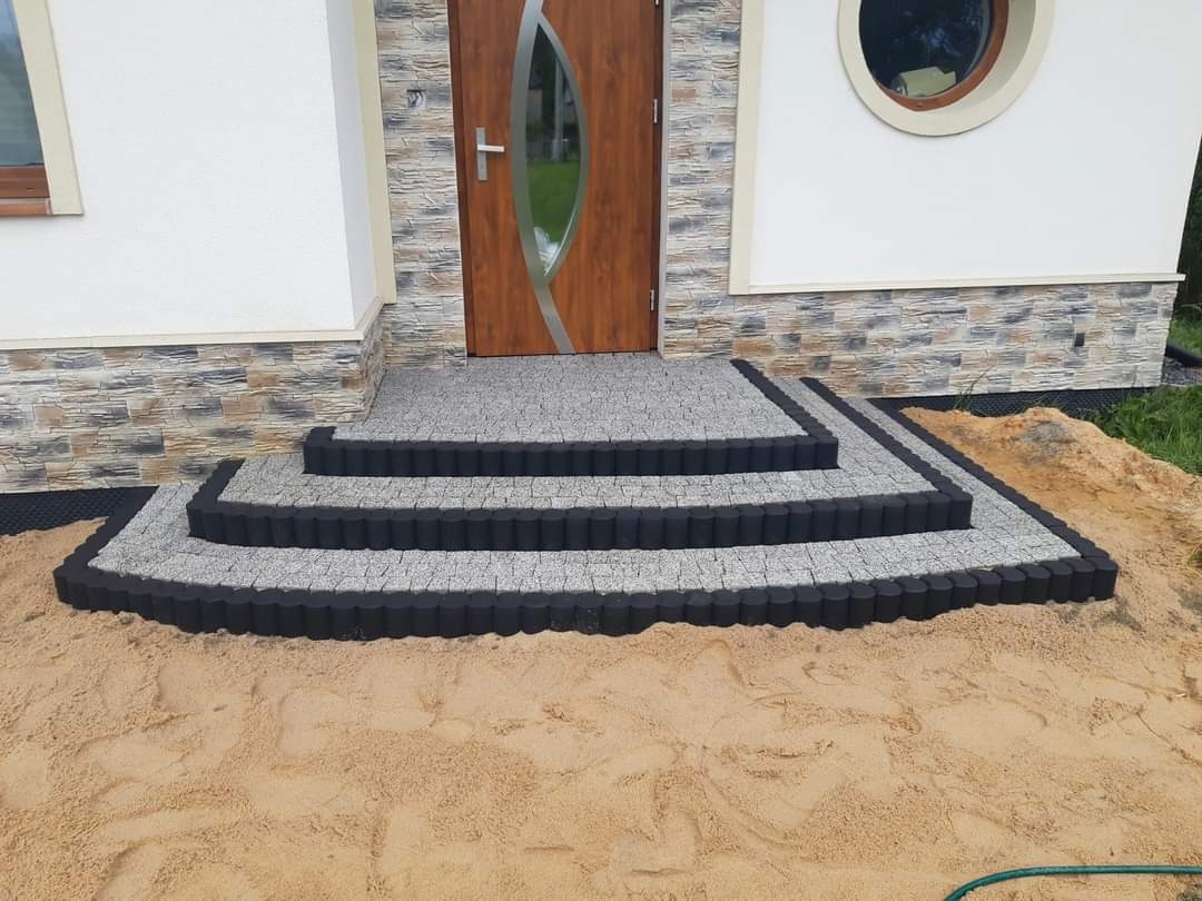 Schody wejściowe do domu wyłożone szarą kostką z czarnymi obrzeżami, otoczone piaskiem.