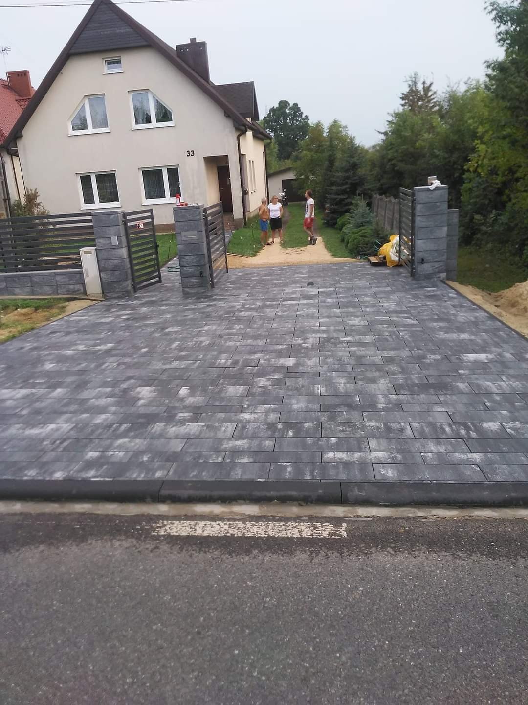 Szeroki podjazd z szarej kostki brukowej, zakończony czarnym obrzeżem, prowadzący do domu z szarą, nowoczesną bramą w tle, widoczni ludzie.