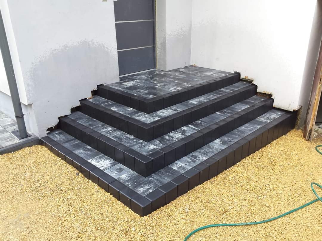 Schody wejściowe wyłożone szarą kostką brukową z czarnym obramowaniem, otoczone drobnym żwirem, widoczne niedociągnięcia w wykończeniu przy ścianie budynku.