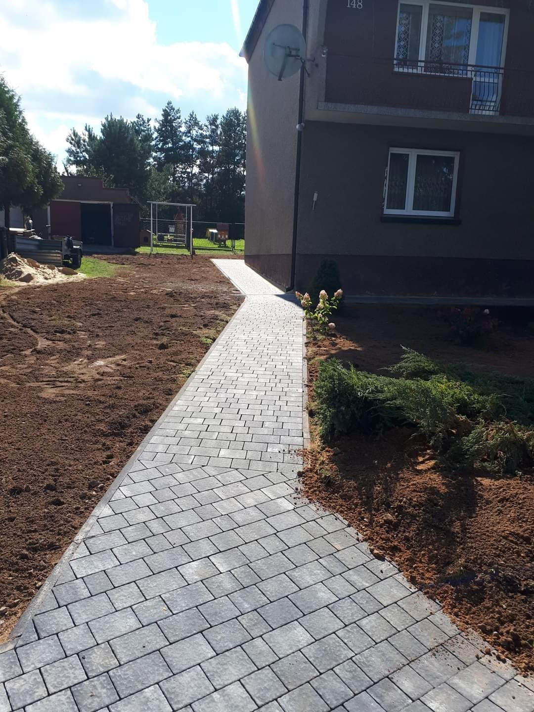 Ścieżka z szarej kostki prowadząca do domu z numerem 148, widoczna antena satelitarna, świeżo obsypana ziemią rabata po prawej stronie.