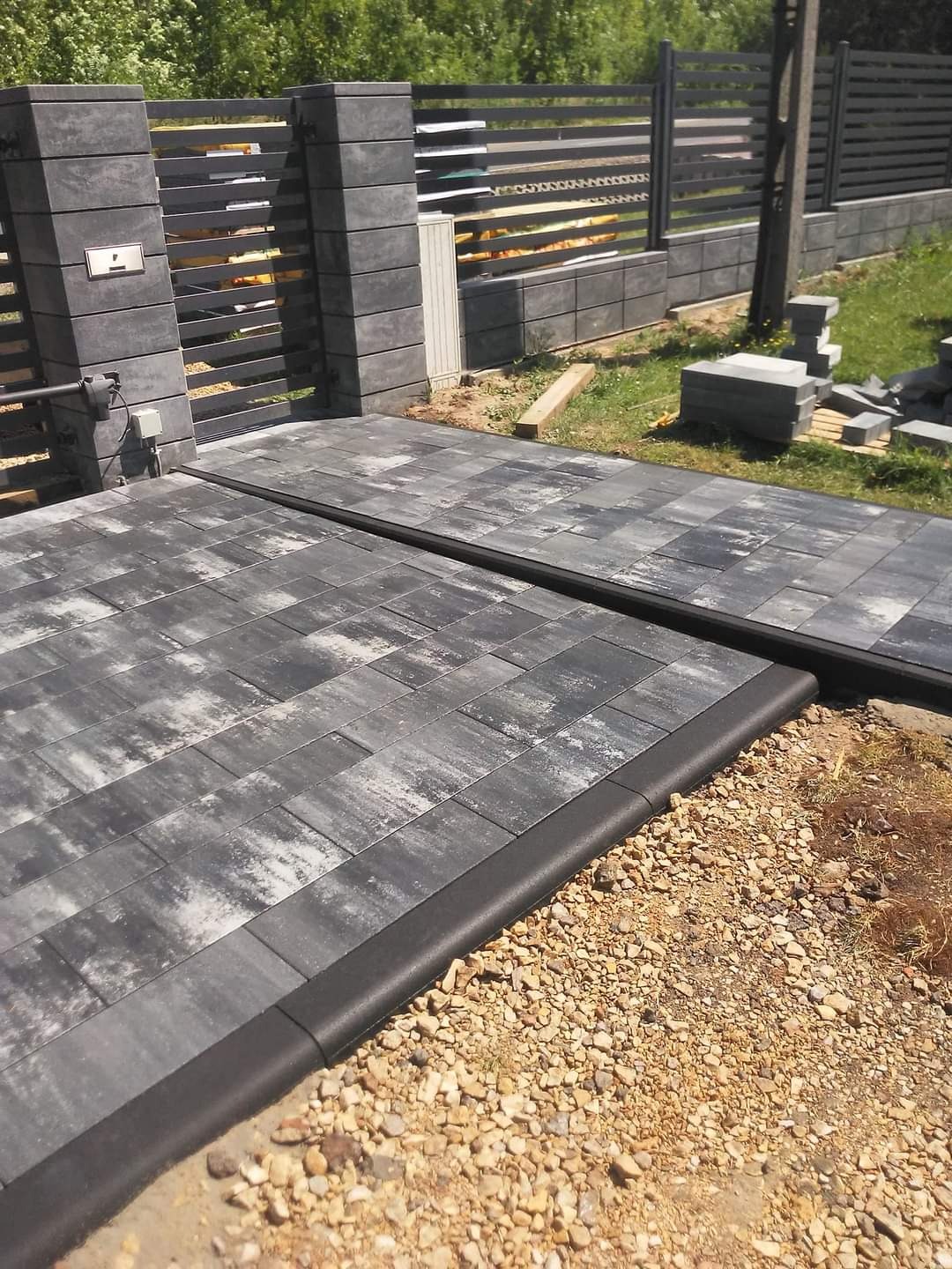 Automatyczna brama wjazdowa z szarej kostki brukowej, obrzeża z czarnym wykończeniem, pasujący szary płot panelowy w tle, widoczne niedokończone prace i stos kostki.