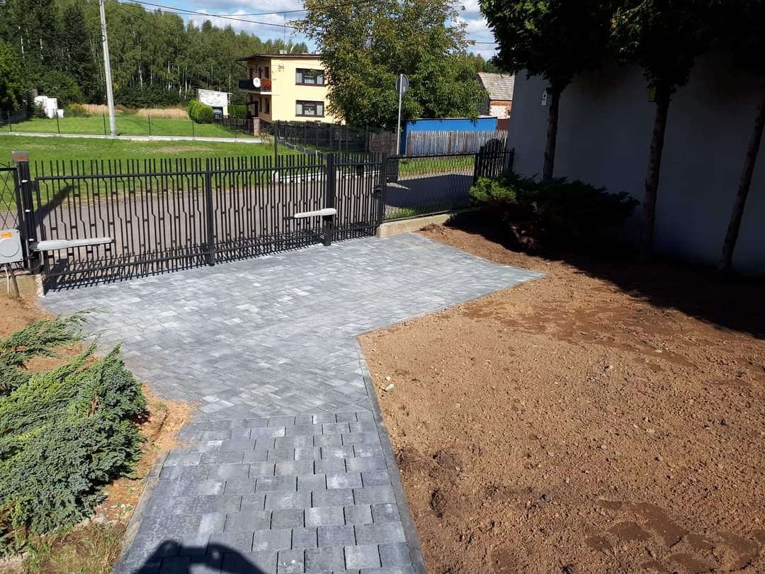 Nowo ułożona, szara kostka brukowa prowadząca do nowoczesnej, czarnej bramy wjazdowej z automatyką, obok przygotowany teren pod trawnik.