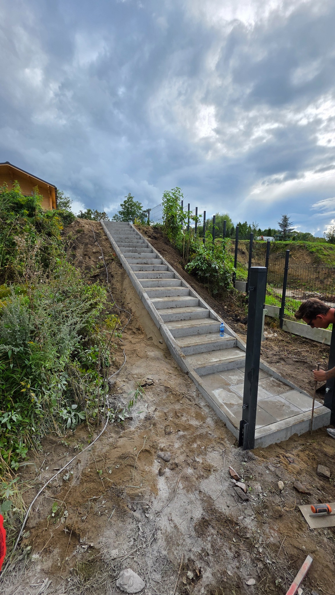 Budowa schodów z kostki brukowej prowadzących na wzniesienie, w Nowogródku Pomorskim. Widoczny fragment ogrodzenia panelowego w tle i pracownik w trakcie montażu.