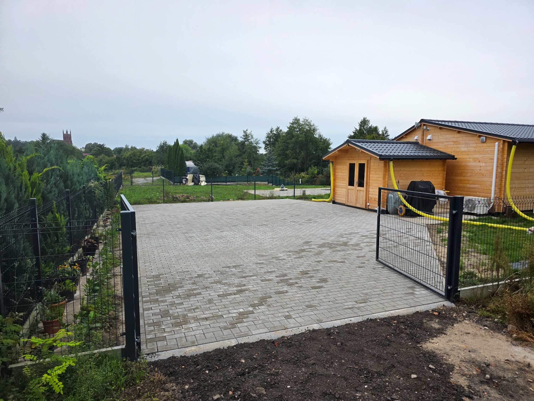 Ułożona kostka brukowa na podjeździe z otwartą bramą panelową, widok na drewniany domek i zieleń w tle. Szara kostka, czarne ogrodzenie.