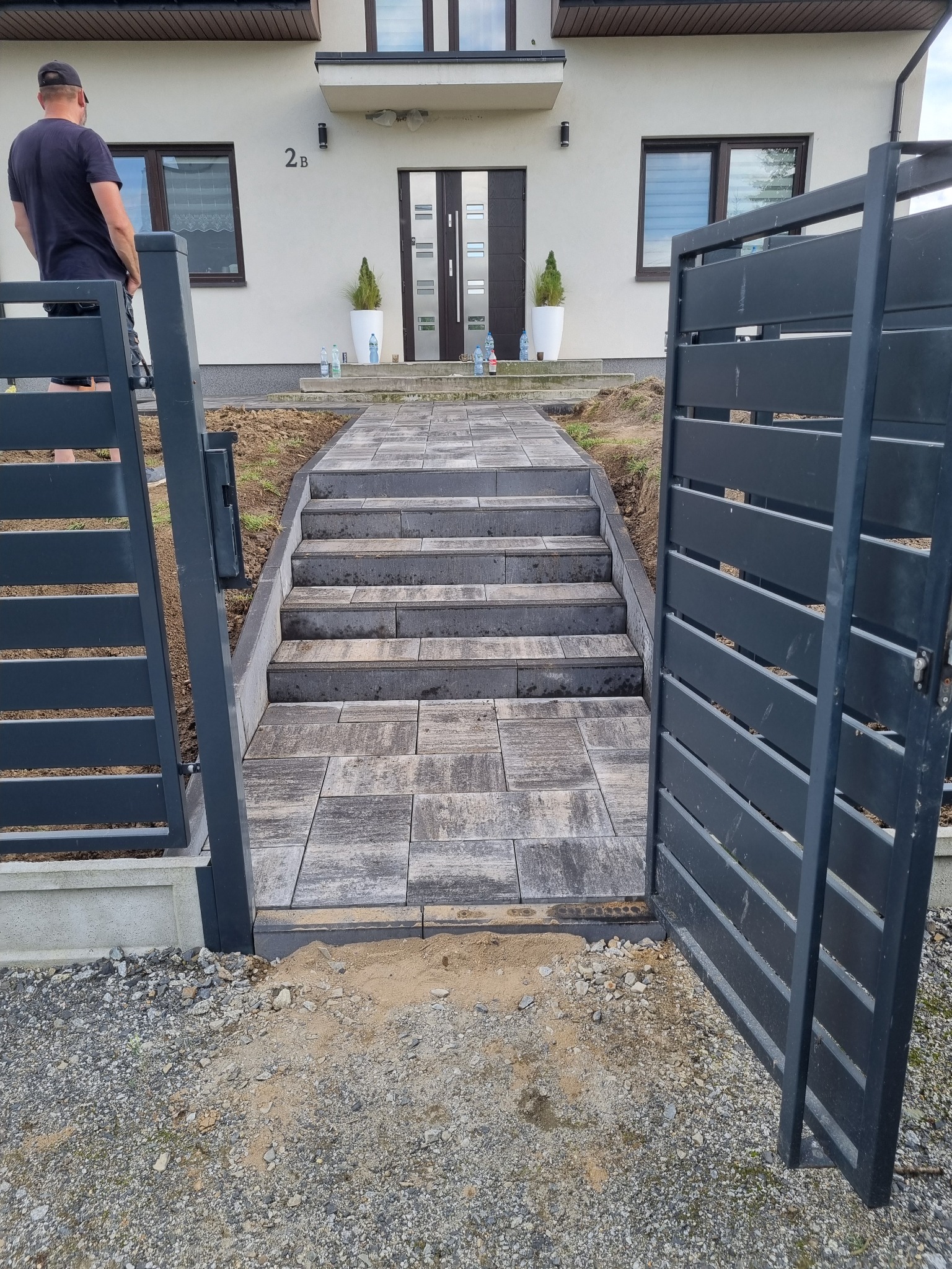 Nowoczesny podjazd z kostki brukowej prowadzący do domu, z otwartą, grafitową bramką panelową po prawej. Widoczny fragment osoby po lewej stronie kadru.