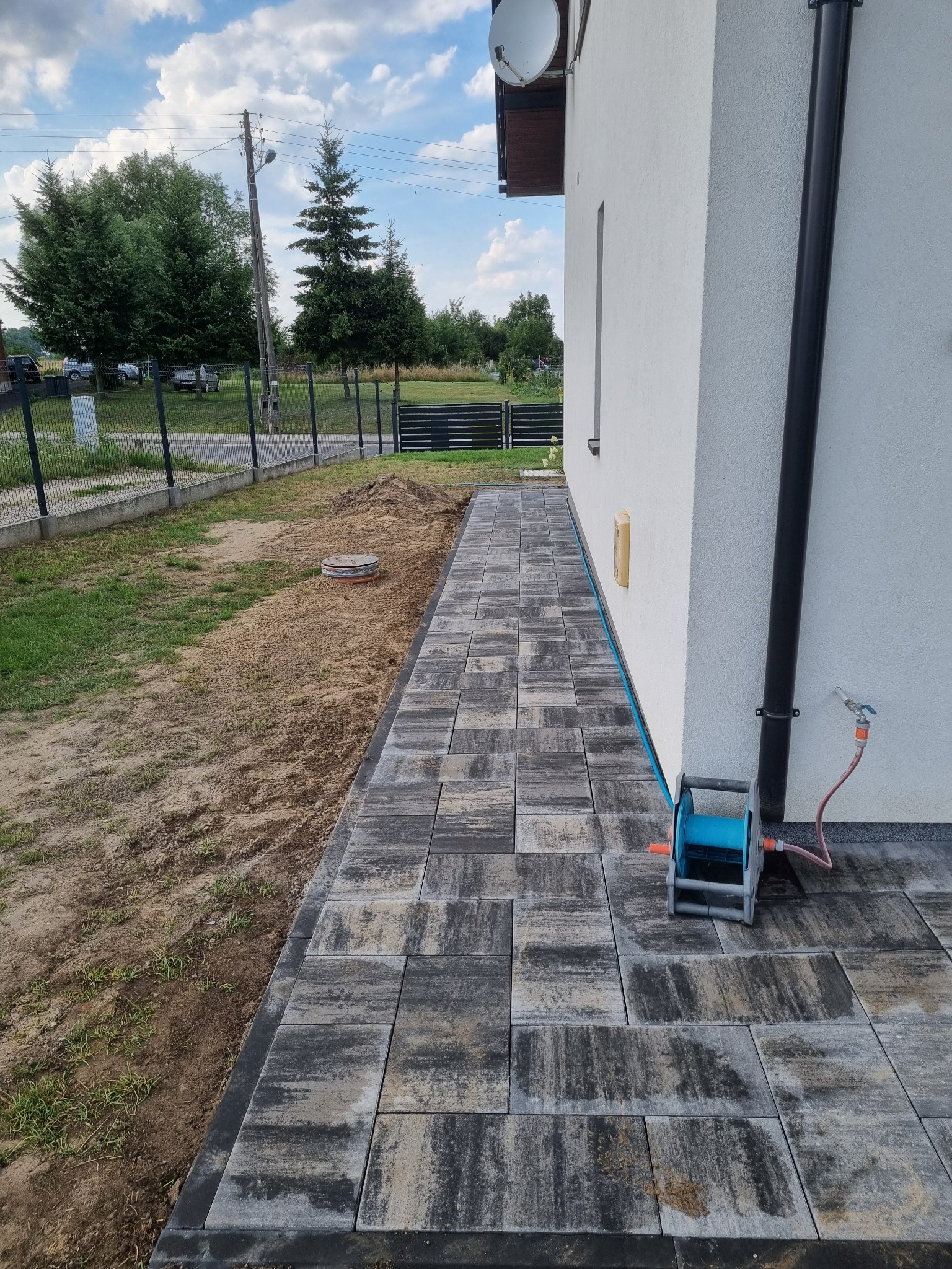 Ułożona ścieżka z szarej kostki brukowej obok białego domu. Widoczny trawnik z ogrodzeniem w tle i zwinięty wąż ogrodowy przy ścianie.