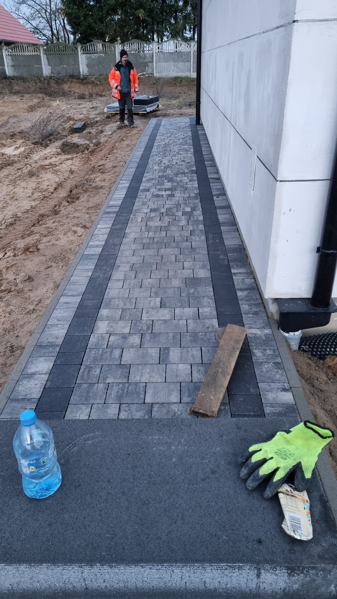 Ułożona ścieżka z kostki brukowej w odcieniach szarości, z ciemniejszym obramowaniem, prowadząca wzdłuż budynku. Widoczne narzędzia i materiały budowlane.