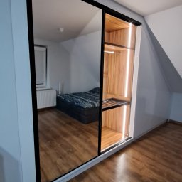 Adrian Paśko ADiMEB - Szafa wnękowa z lustrzanymi drzwiami przesuwnymi, oświetleniem LED wewnątrz i czarną ramą, odbijająca wnętrze pokoju z łóżkiem i oknem.