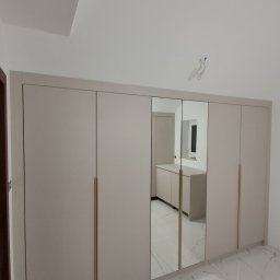 Adrian Paśko ADiMEB - Beżowa szafa wnękowa z lustrzanymi drzwiami i złotymi uchwytami na tle białej marmurowej podłogi, widoczne kable wystające ze ściany.