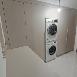 Adrian Paśko ADiMEB - Beżowa zabudowa meblowa z pralką i suszarką w kolumnie, marmurowa podłoga, minimalistyczne wnętrze.