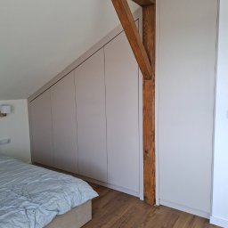 Adrian Paśko ADiMEB - Jasnoszara szafa wnękowa na poddaszu, wkomponowana w skos dachu, z widoczną drewnianą belką konstrukcyjną i fragmentem łóżka z pościelą.