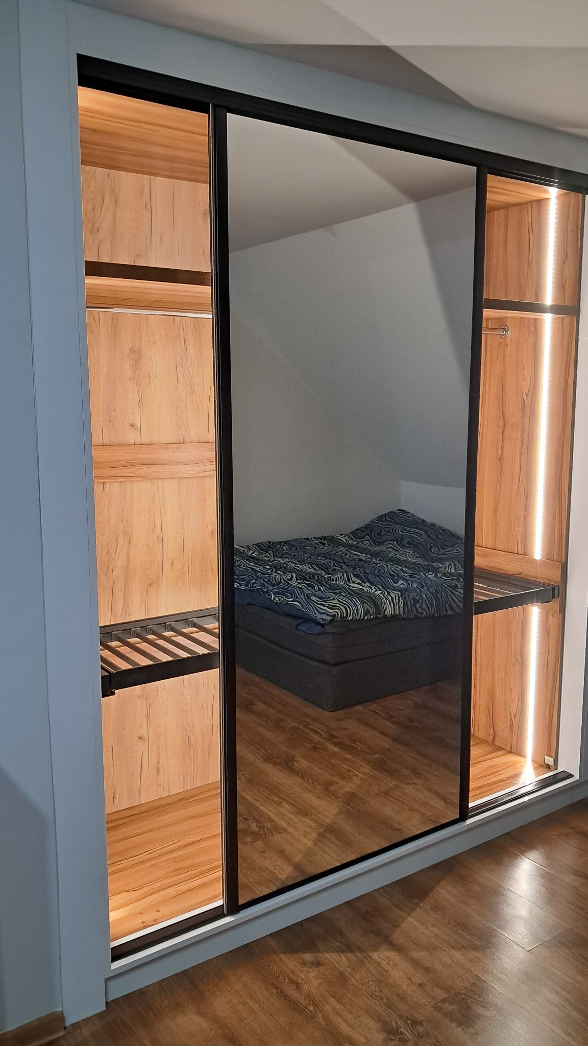 Szafa przesuwna z lustrem odbijającym sypialnię; wnętrze szafy oświetlone listwami LED, wykończone drewnopodobną okleiną.