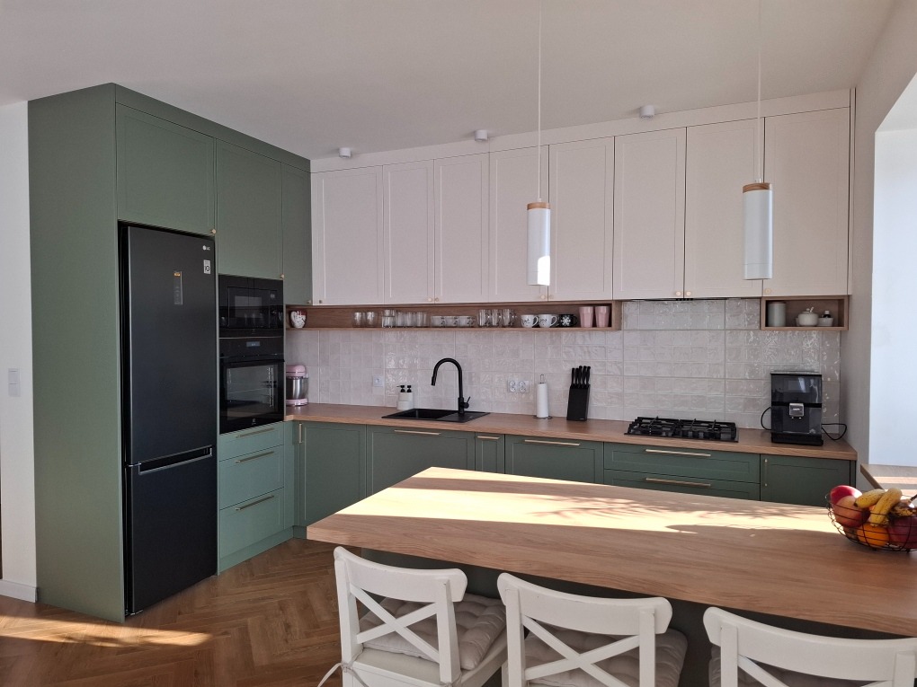 Nowoczesna kuchnia z zielonymi i białymi szafkami, drewnianym blatem i czarnym sprzętem AGD, oświetlona wiszącymi lampami z drewnianymi akcentami.