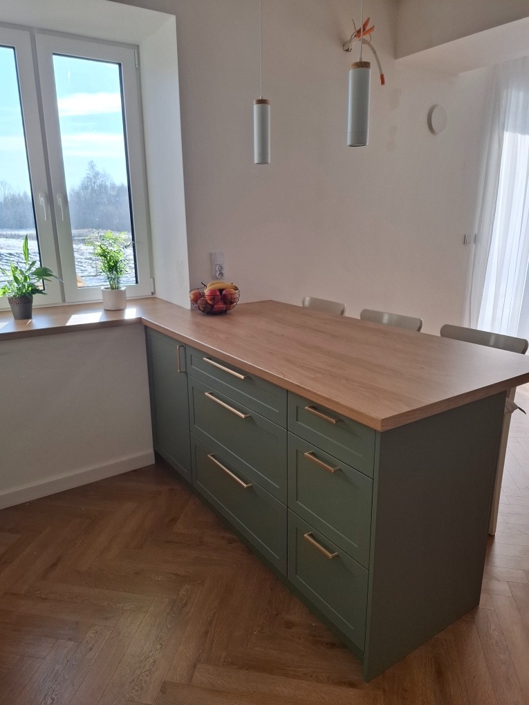 Wyspa kuchenna z zielonymi szafkami i drewnianym blatem, widok na okno z roślinami, parkiet w jodełkę na podłodze, minimalistyczne lampy wiszące.