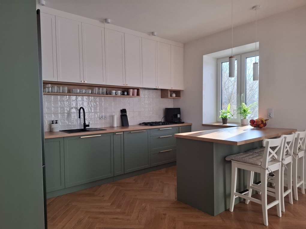 Nowoczesna kuchnia z zielonymi szafkami dolnymi i białymi górnymi, drewniany blat, wyspa kuchenna z krzesłami barowymi, parkiet w jodełkę, oświetlenie punktowe.