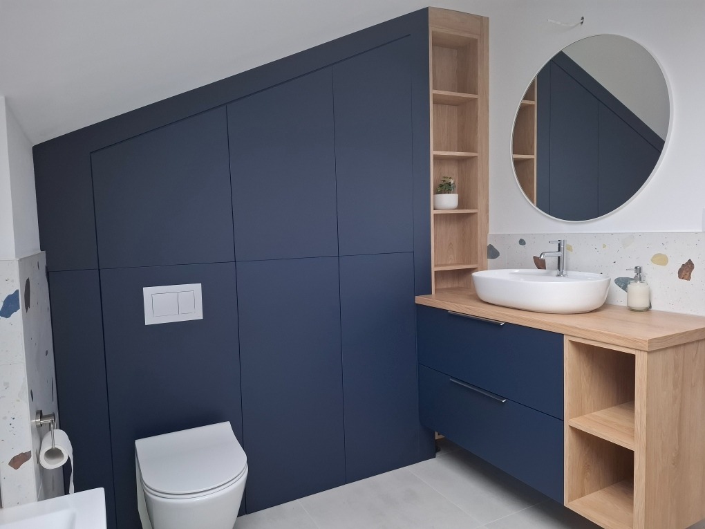 Niebieska szafa na wymiar zabudowana pod skosem w łazience, obok umywalka nablatowa z drewnianym blatem i otwartym regałem, okrągłe lustro.