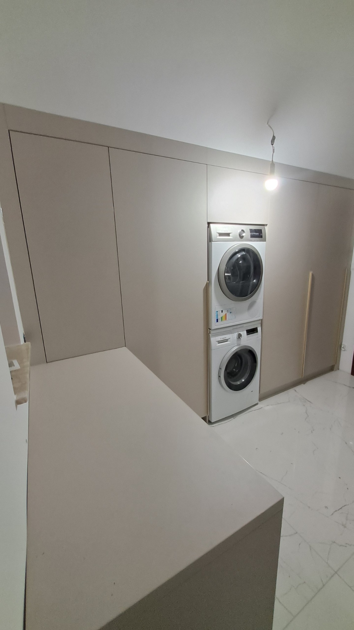 Beżowa zabudowa meblowa z pralką i suszarką w kolumnie, marmurowa podłoga, minimalistyczne wnętrze.