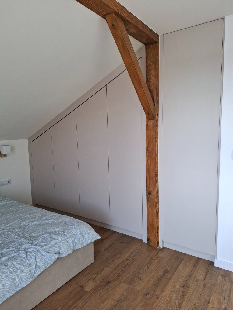 Jasnoszara szafa wnękowa na poddaszu, wkomponowana w skos dachu, z widoczną drewnianą belką konstrukcyjną i fragmentem łóżka z pościelą.