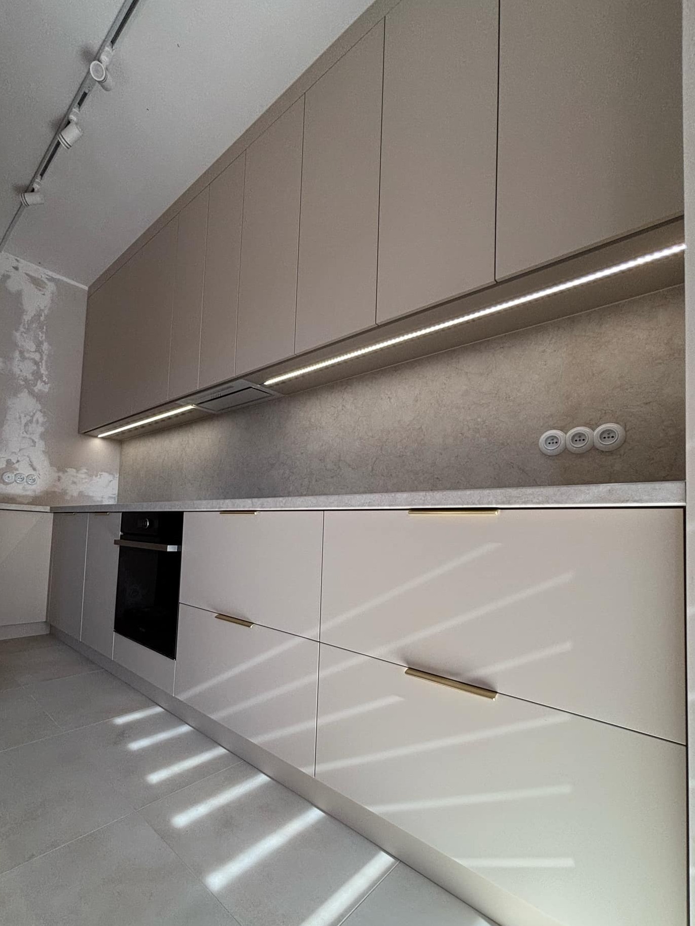 Jasna kuchnia na wymiar z oświetleniem LED pod szafkami wiszącymi i złotymi uchwytami, widoczny fragment ściany przed wykończeniem.