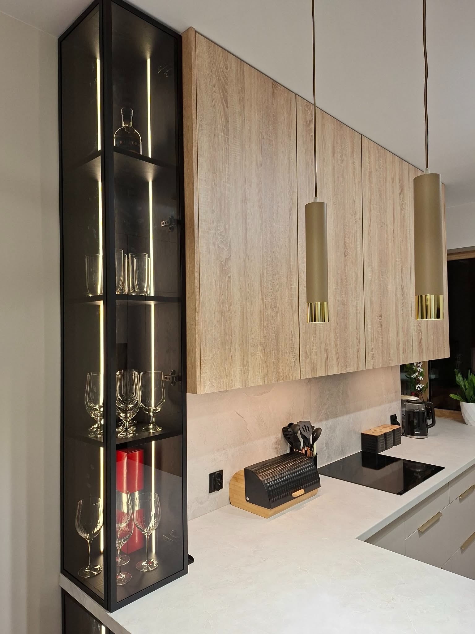Nowoczesna kuchnia z drewnianymi szafkami wiszącymi i przeszkloną witryną z podświetleniem LED, eksponującą szkło i dekoracje. Minimalistyczne lampy wiszące.