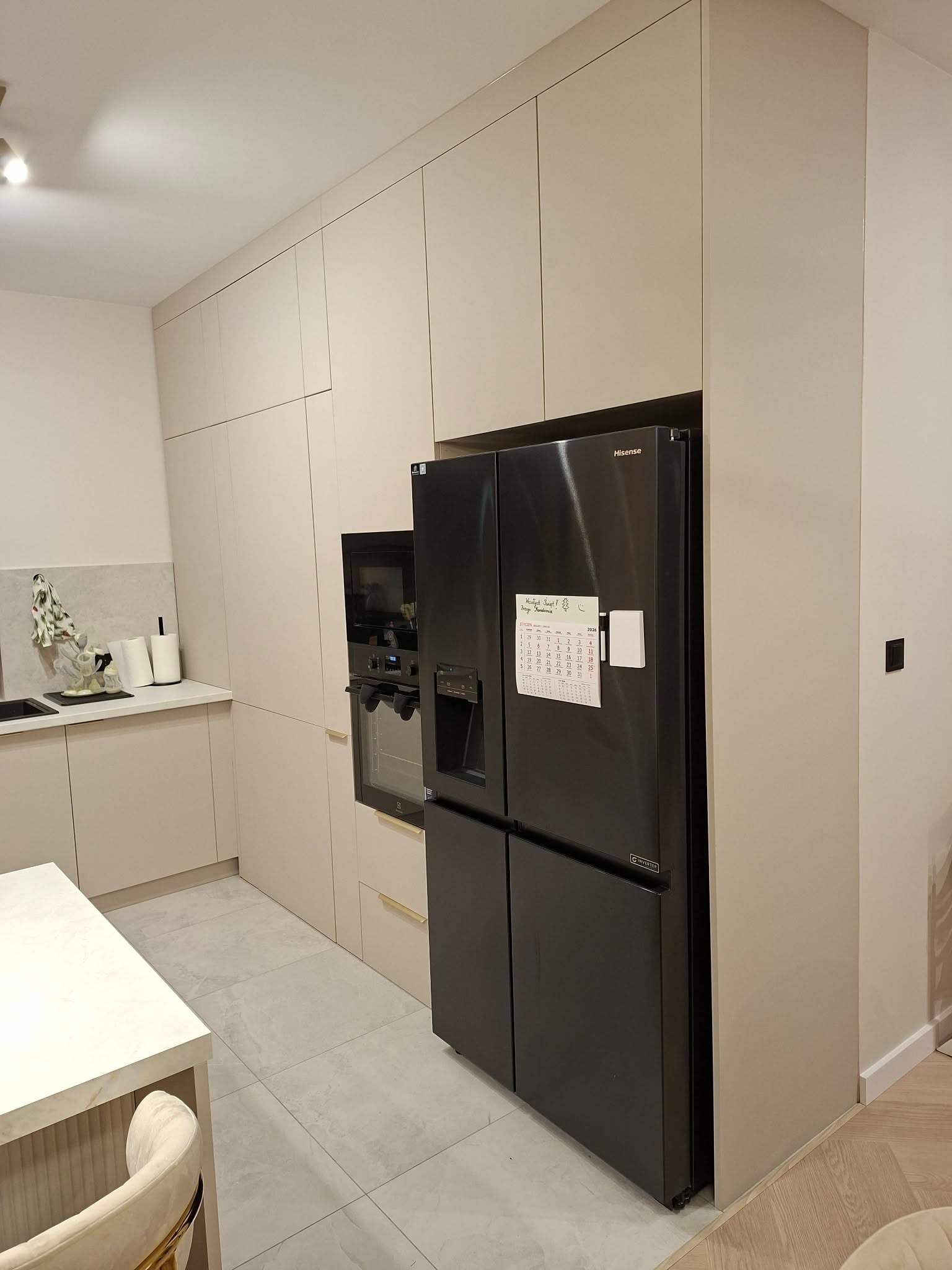 Jasna kuchnia z zabudową na wymiar, czarna lodówka Hisense z kalendarzem na drzwiach, piekarnik w zabudowie i marmurowy blat. Minimalistyczny design.
