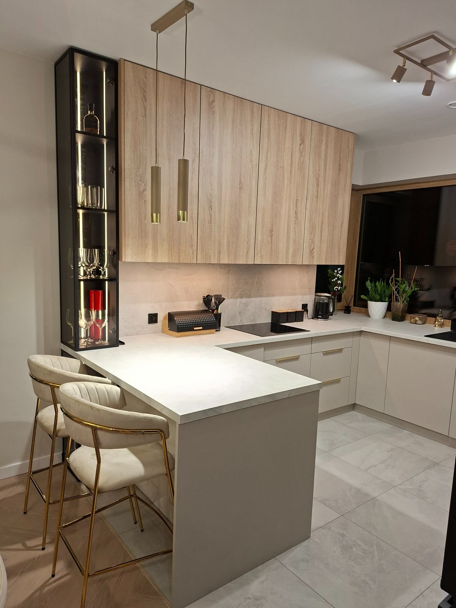Nowoczesna kuchnia z wyspą, drewnianymi szafkami górnymi i podświetlaną witryną. Dwa krzesła barowe z beżową tapicerką i złotymi nogami. Minimalistyczne lampy wiszące.