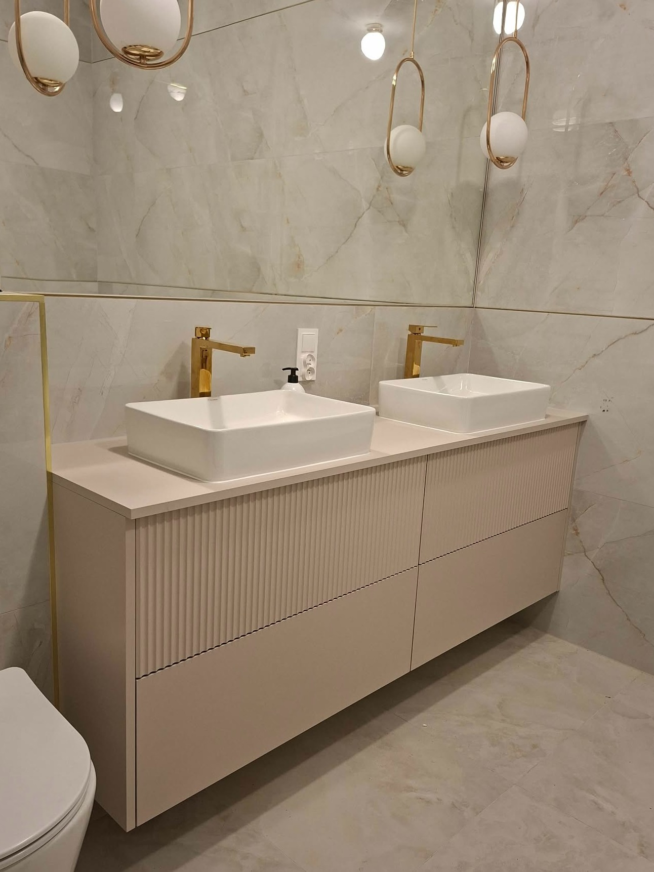 Łazienka z beżową szafką pod umywalki, złotymi bateriami i marmurowymi płytkami. Uwagę przykuwają wiszące lampy z kulistymi kloszami.