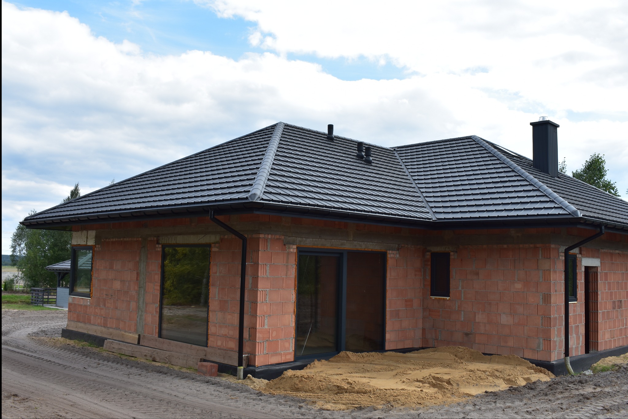 Nowo wybudowany dom w stanie surowym zamkniętym z ciemnoszarą dachówką ceramiczną i czarnymi rynnami, widoczne obróbki blacharskie i komin.