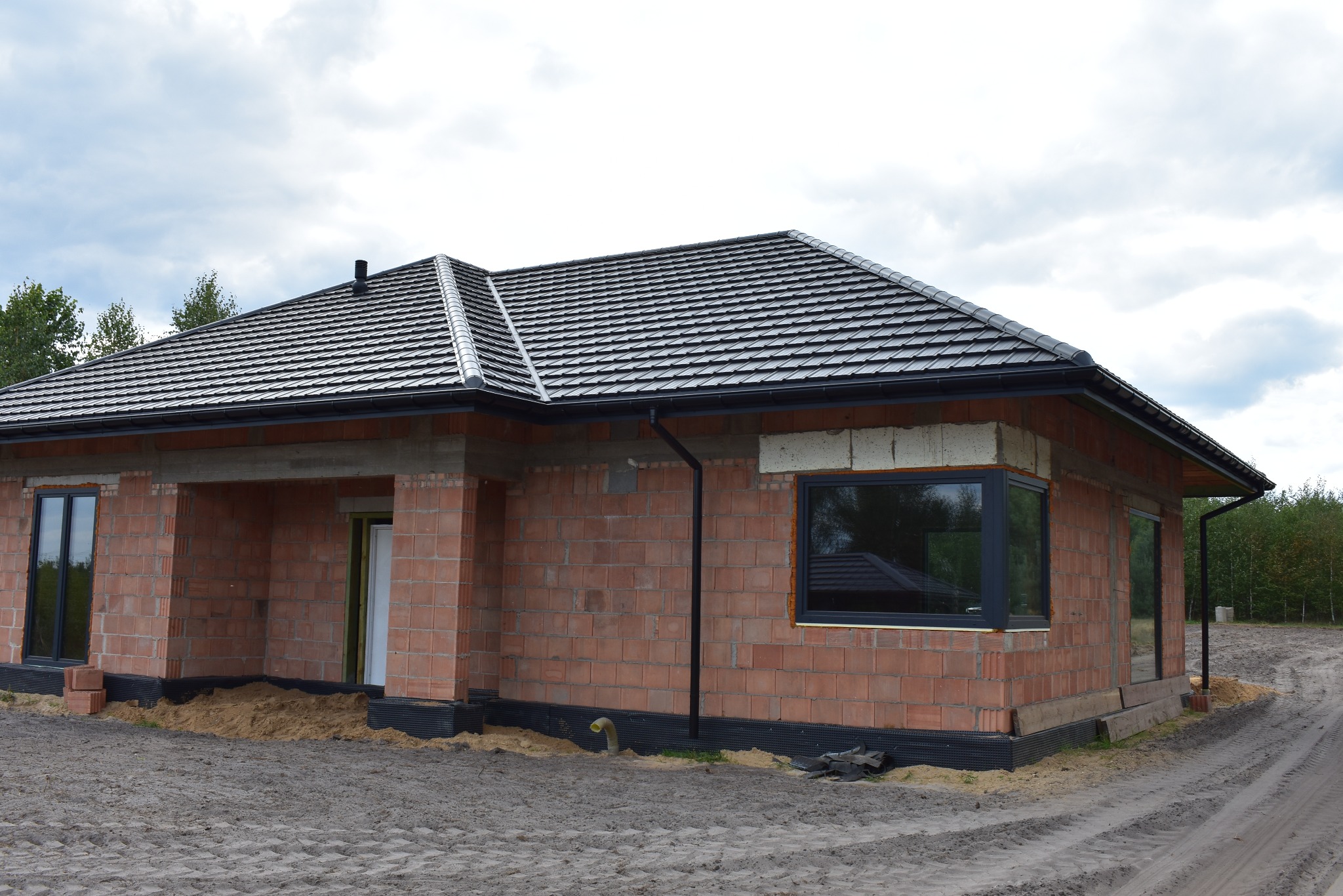 Surowy budynek jednorodzinny z ciemną dachówką ceramiczną i czarnymi rynnami, w trakcie budowy, widoczne cegły i piasek wokół fundamentów.