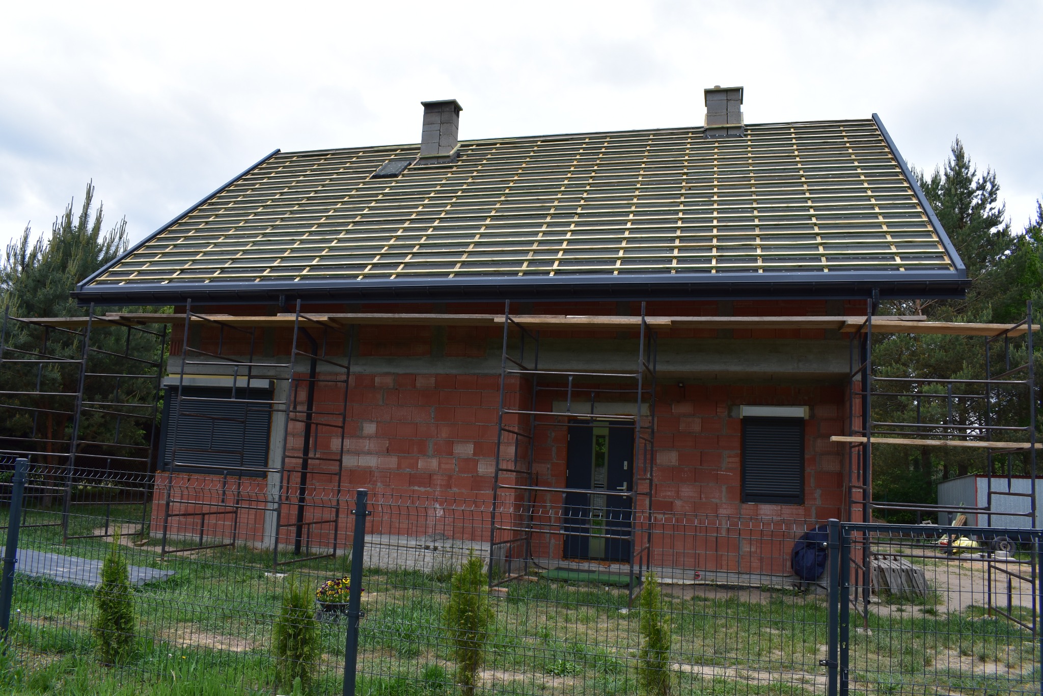 Dom w trakcie budowy, z widoczną konstrukcją drewnianą pod przyszłe pokrycie dachowe, szare rynny i kominy z cegły, otoczony rusztowaniami i ogrodzeniem z siatki.