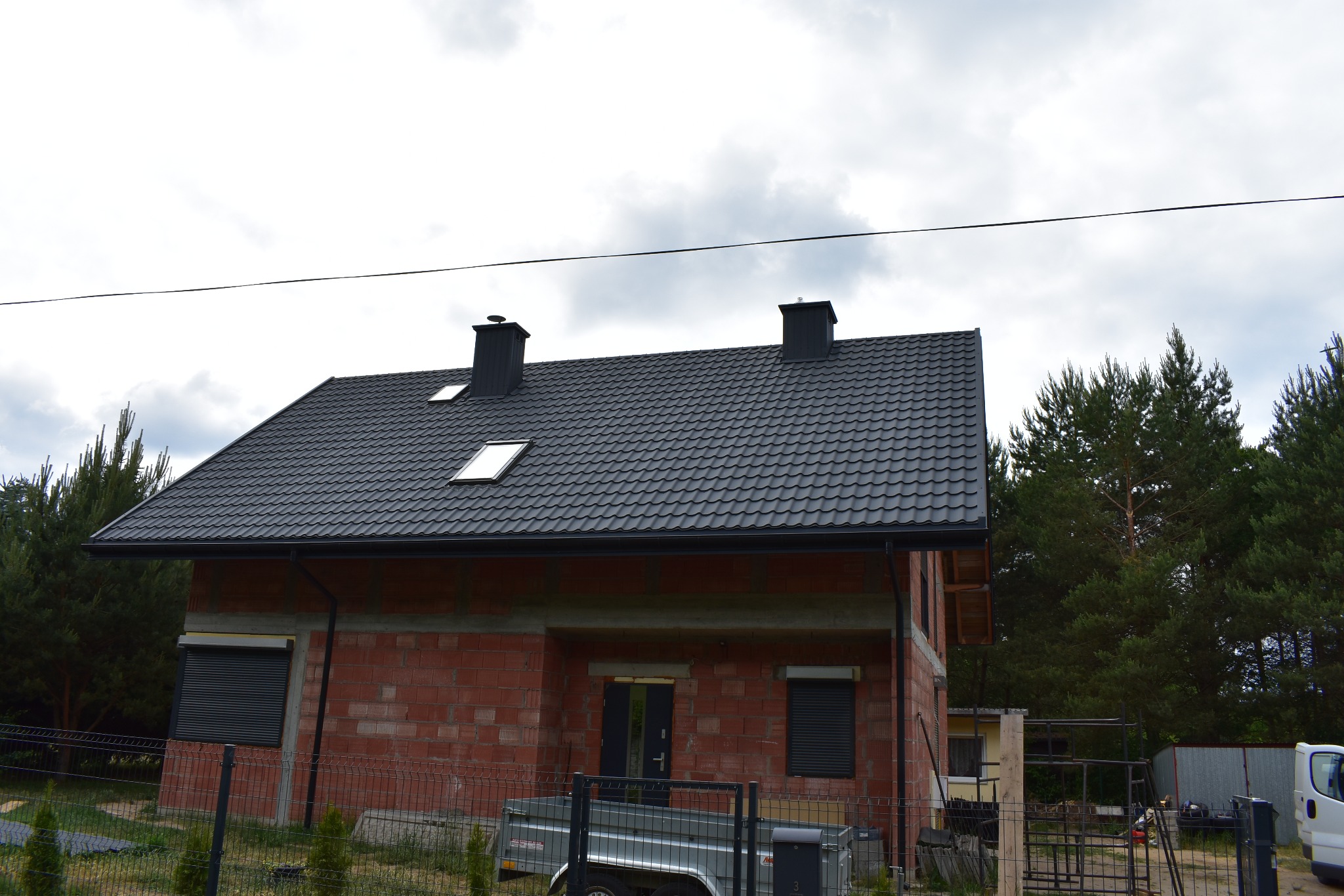 Dom w trakcie budowy z ciemnoszarym dachem z dachówki modułowej, dwoma kominami i oknami dachowymi, widoczny z boku, z przyczepką i ogrodzeniem na pierwszym planie.