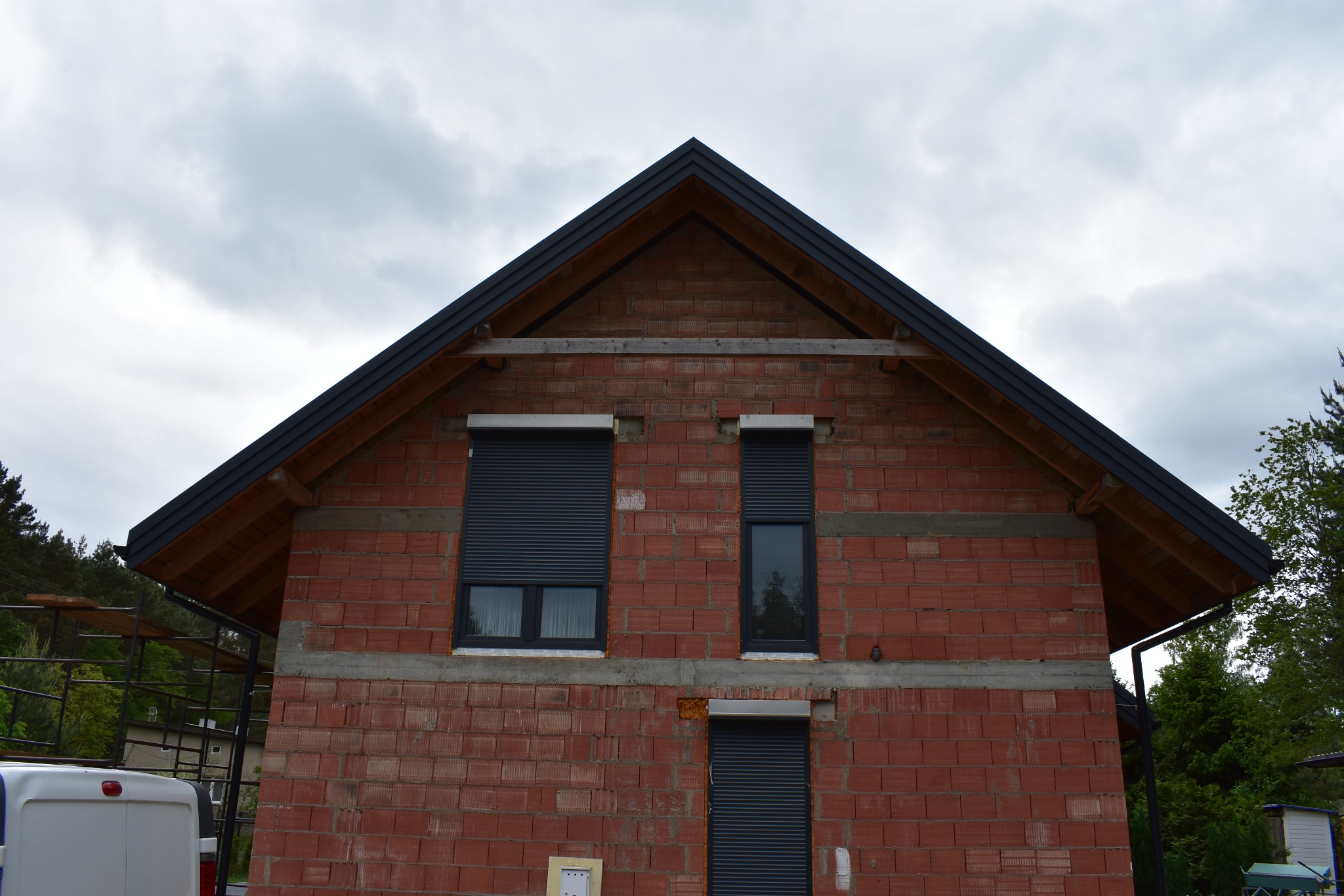 Surowy stan budynku z czerwonej cegły z zamontowanymi roletami zewnętrznymi i częściowo wykończonym dachem z czarną blachą, widoczna drewniana konstrukcja dachu.