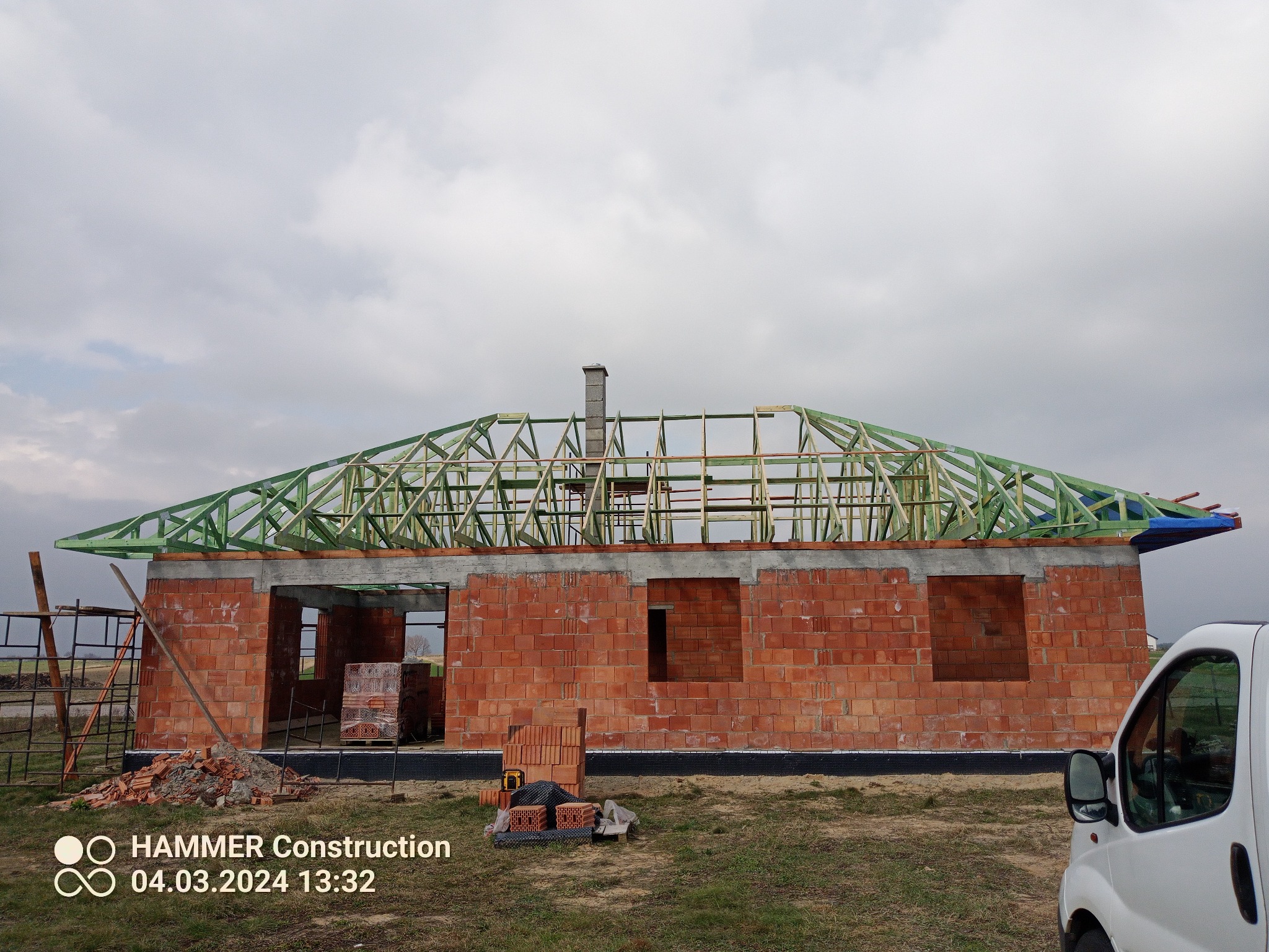 Budowa domu jednorodzinnego w trakcie wznoszenia, widoczna konstrukcja ścian z cegły oraz zielona drewniana konstrukcja dachu, częściowo pokryta niebieską folią, na tle pochmurnego nieba.