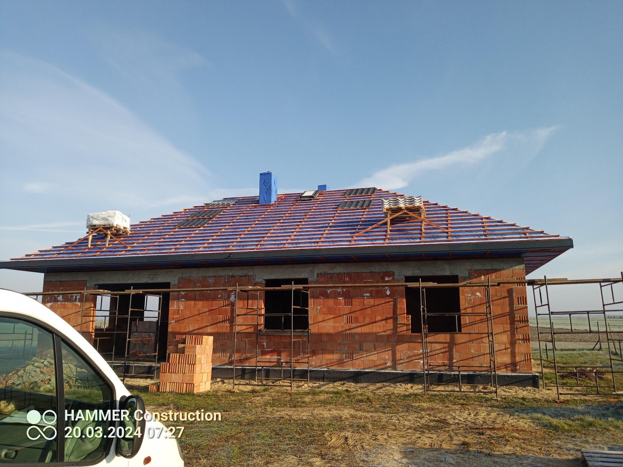 Budowa domu jednorodzinnego w trakcie, widoczna konstrukcja dachu z łatami i częściowo ułożoną dachówką, ściany z czerwonej cegły, rusztowania i stos cegieł przed budynkiem, fragment białego...