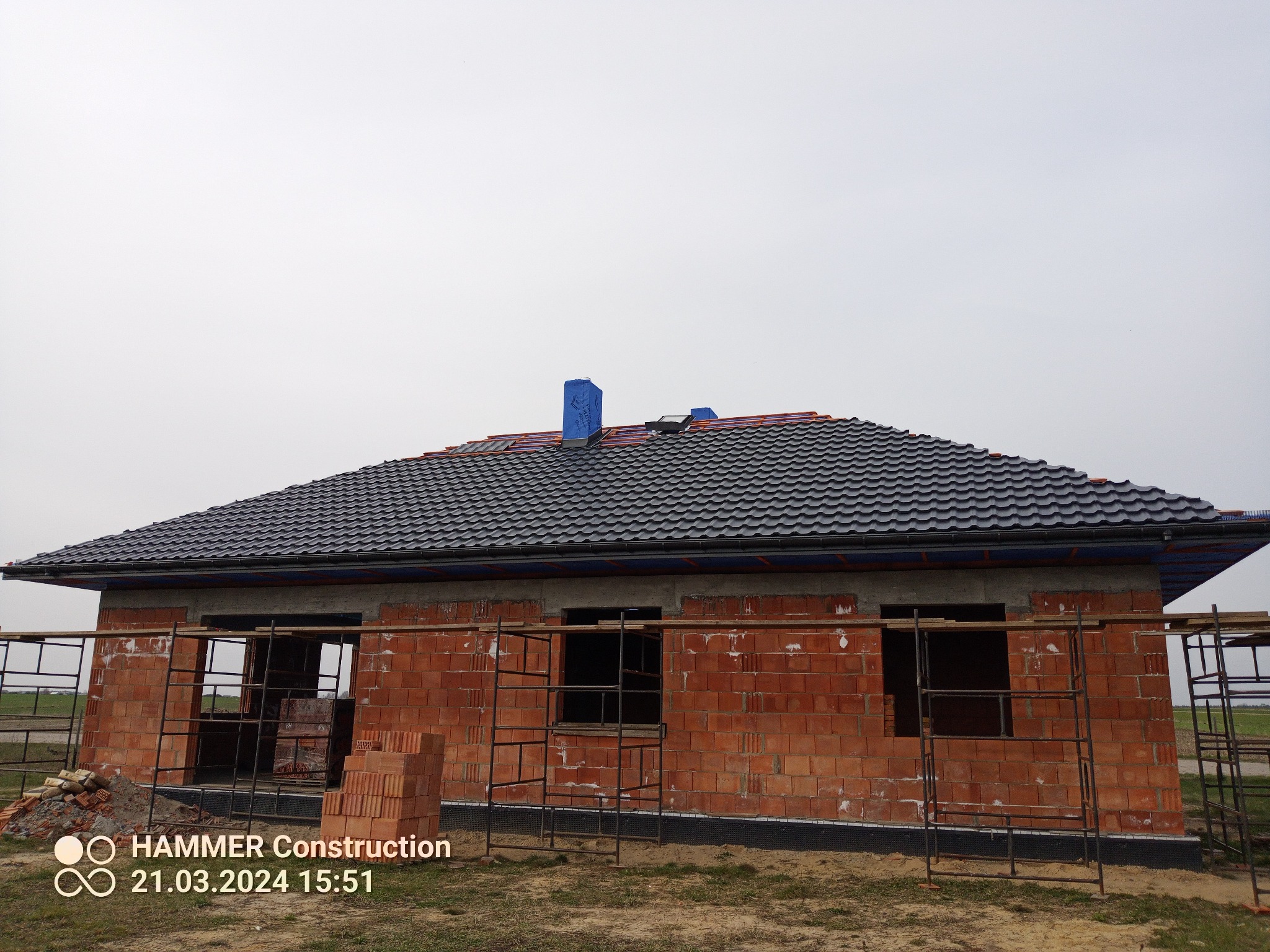 Niedokończony dom jednorodzinny z ciemną dachówką, widoczne rusztowania i materiały budowlane, niebieskie zabezpieczenie komina. Zdjęcie wykonane w pochmurny dzień.