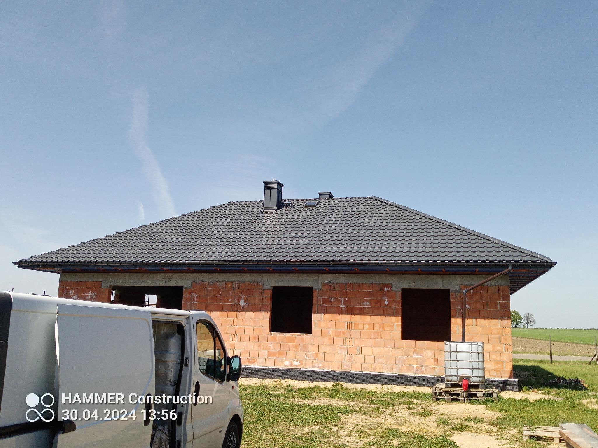 Dom w trakcie budowy z ciemnoszarą dachówką, widoczny fragment białego samochodu dostawczego i zbiornik na wodę obok budynku.
