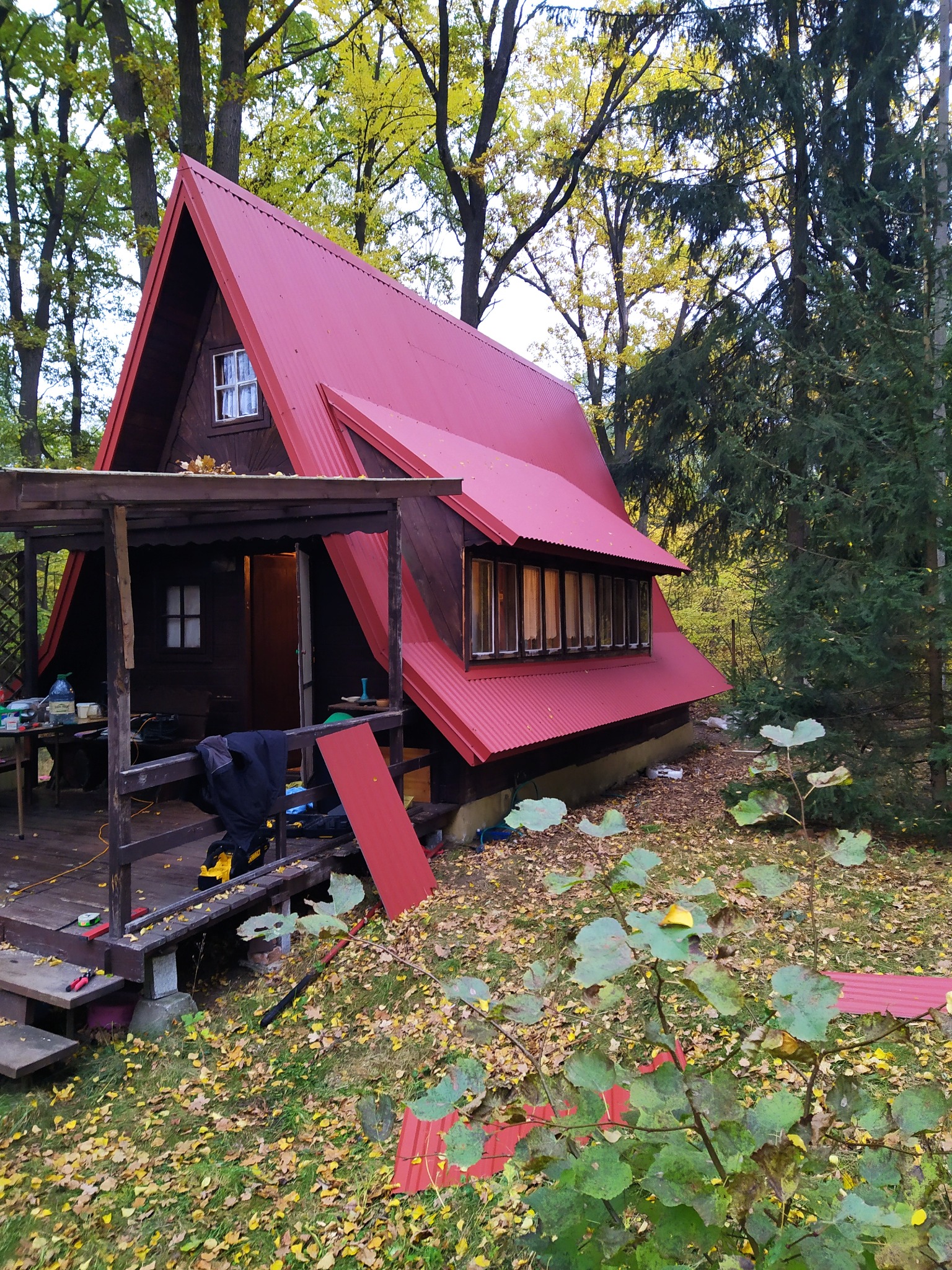 Domek letniskowy typu A-frame z czerwonym blaszanym dachem w otoczeniu drzew, z widocznymi narzędziami i elementami blachy na ziemi, sugerującymi prace dekarskie w toku.