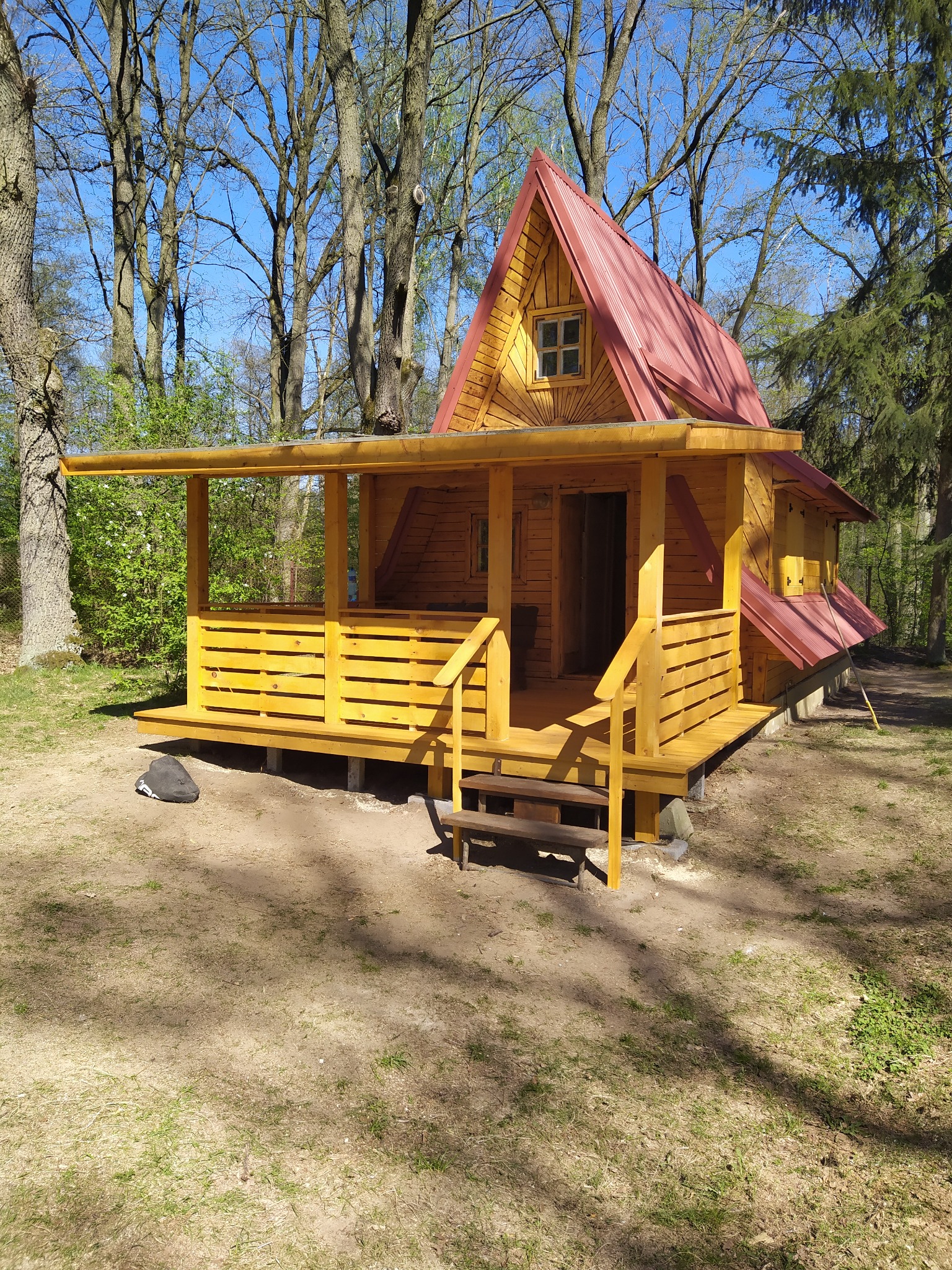 Mały, drewniany domek z czerwonym, metalowym dachem w lesie, z widoczną werandą i schodami wejściowymi, świeżo pomalowany na żółto, z torbą leżącą obok.