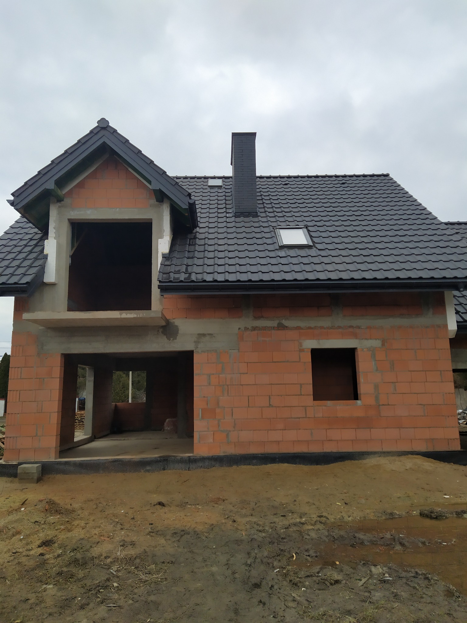 Surowy stan domu jednorodzinnego z czerwoną cegłą, czarną dachówką i kominem, widoczne okno dachowe, brak okien i drzwi, teren wokół domu niezagospodarowany, pochmurne niebo.
