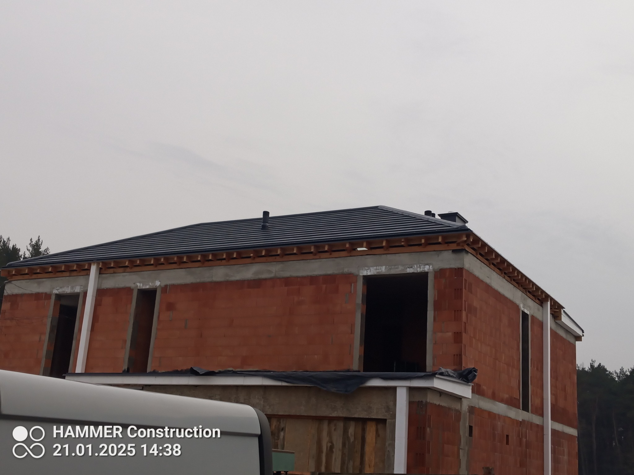 Dom w trakcie budowy z częściowo ukończonym dachem z czarnej dachówki, widoczne niezabudowane otwory okienne i drzwiowe, oraz fragment samochodu z logiem firmy budowlanej.