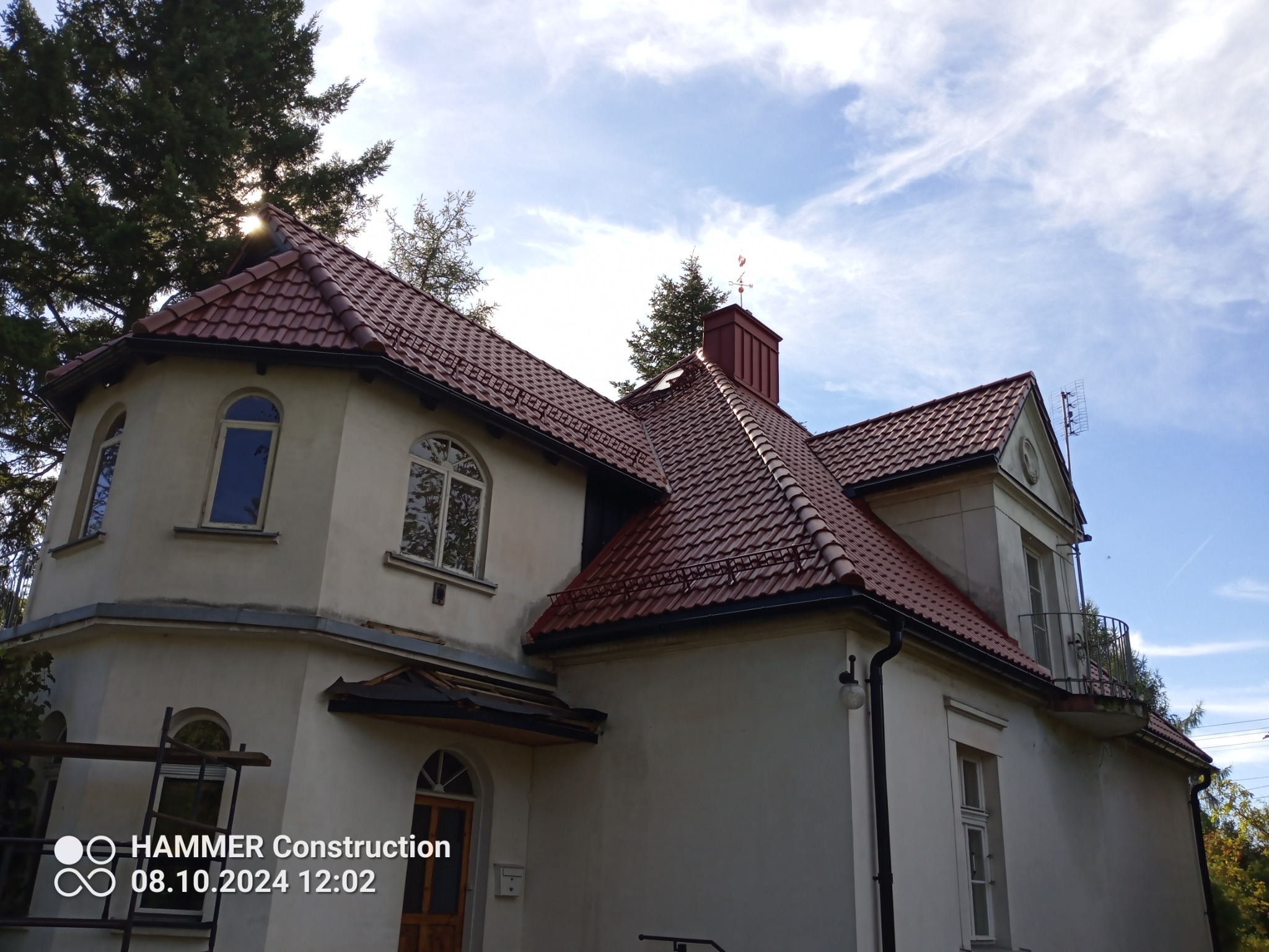 Dwupiętrowy dom z nową, czerwoną dachówką ceramiczną, widoczny komin z wiatrowskazem, na tle błękitnego nieba z chmurami i drzew.