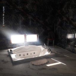 R.M.S. BUD Robert Mirociński - Biała wanna z hydromasażem na tle ciemnych marmurowych ścian, oświetlona promieniami słońca wpadającymi przez okno.