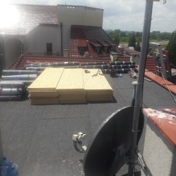 R.M.S. BUD Robert Mirociński - Przygotowanie do krycia dachu: rolki papy, płyty OSB i narzędzia na płaskim dachu budynku z widocznymi elementami wentylacji i anteną satelitarną.