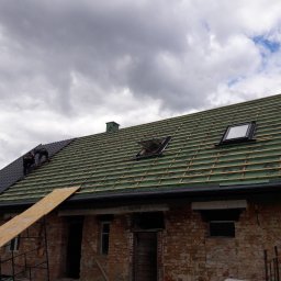 R.M.S. BUD Robert Mirociński - Częściowo pokryty dach z dwoma oknami dachowymi na tle zachmurzonego nieba, widoczni dekarze pracujący na dachu ceglanego budynku.