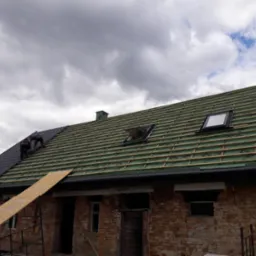 Częściowo pokryty dach z dwoma oknami dachowymi na tle zachmurzonego nieba, widoczni dekarze pracujący na dachu ceglanego budynku.