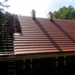 Częściowo pokryty dach z brązowej dachówki ceramicznej, widoczna więźba dachowa i kominy, drabina oparta o budynek