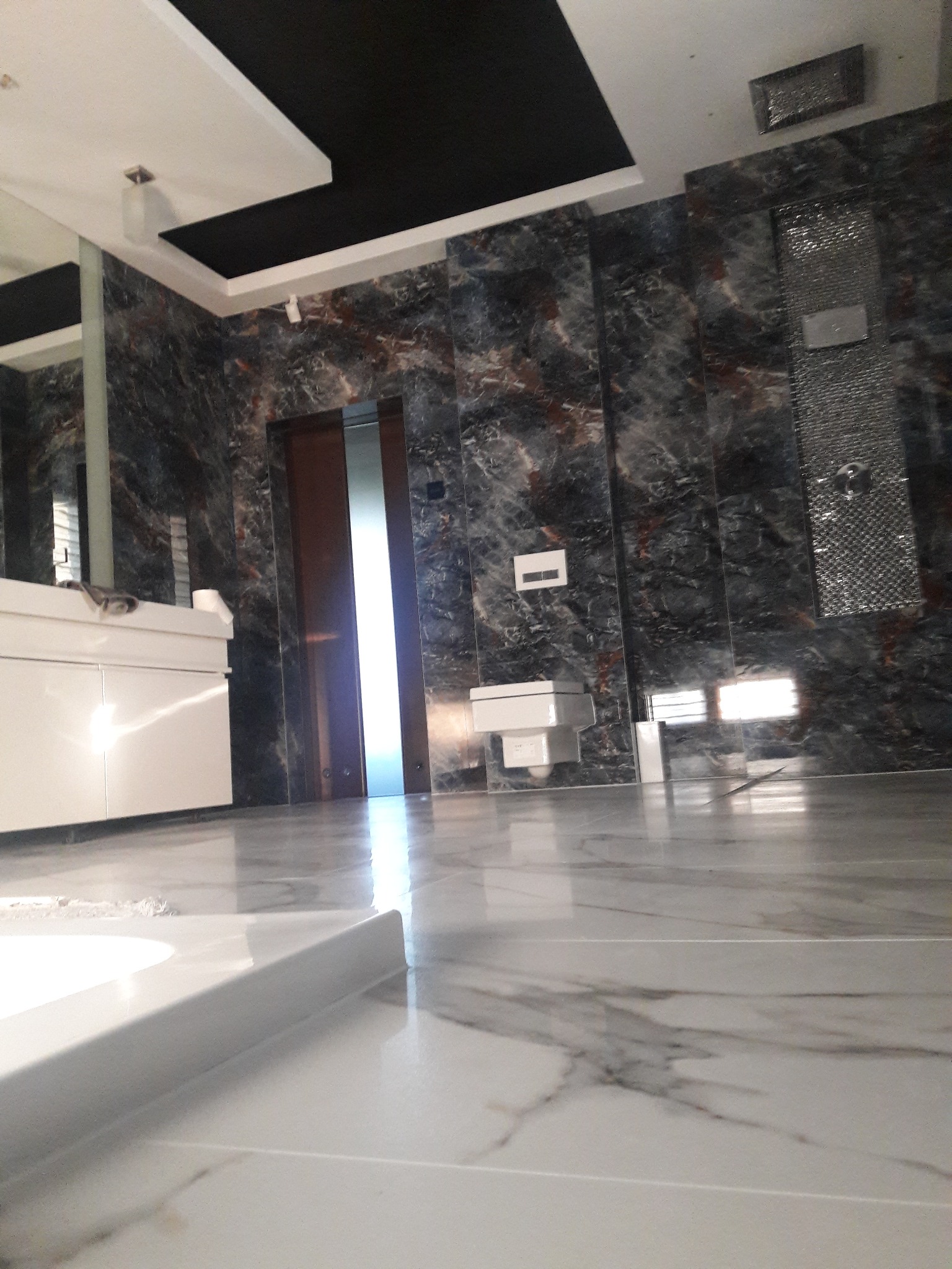 Wykończona łazienka z marmurowymi ścianami i podłogą, białą armaturą i czarnym sufitem podwieszanym, widok z poziomu podłogi.